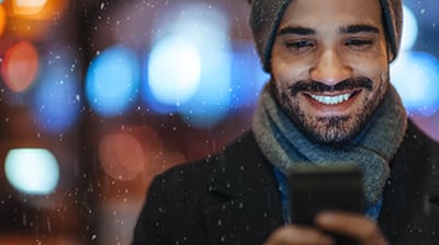 Man dressed in winter clothes smiling as he looks at his phone while it snows