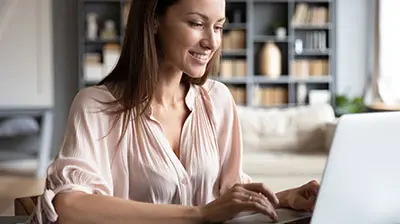A woman in a light pink blouse,  smiling while using a laptop 