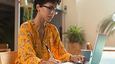 A woman wearing an orange blouse, sitting at a table and using her laptop while holding a pen in one hand 