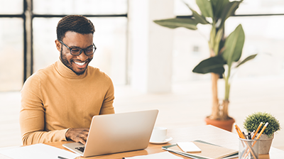 A man sitting at his desk and smiling while using a laptop 